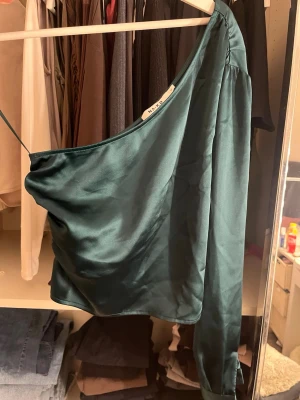 Mörkgrön oneshoulder topp från NA-KD - Snygg mörkgrön oneshoulder topp från NA-KD i glansigt satinliknande material. Toppen har en lång ärm på ena sidan och ett smalt axelband på andra, vilket ger en unik och trendig look. Perfekt för dig som vill sticka ut lite extra.