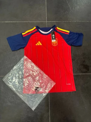 Spaniens matchtröja Adidas röd/blå - Säljer en officiell matchtröja från Spaniens landslag, designad av Adidas. Tröjan är röd med blå ärmar och gula detaljer på axlarna, samt klubbmärke och Adidas-logga på bröstet. Tillverkad i lätt och ventilerande material, perfekt för fotbollsträning eller match.