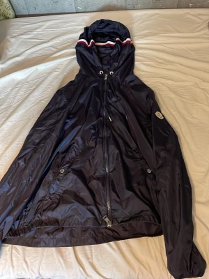Mörkblå vindjacka från Moncler - Snygg mörkblå vindjacka från Moncler med huva och dragkedja framtill. Jackan har vita och röda detaljer på huvan, två fickor med knappar och elastisk nederkant. Tillverkad i glansigt, lätt material som står emot vind. Perfekt för en stilren look. Säljs på grund av att den har blivit för liten, riktigt bra skick, varsamt använd, priset går att diskutera. Tveka inte höra av dig för minsta fundering✅🤝 