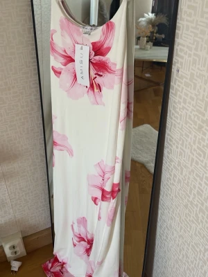 Vit långklänning med rosa blommor Amisu - Långklänning från Amisu i vit med stora rosa blommor. Klänningen är ärmlös och har en rak, flowy passform som ger en avslappnad och somrig vibe. Perfekt för varma dagar och midsommarfirande. Materialet känns mjukt och svalt mot huden.