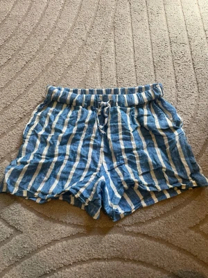 Randiga blåvita shorts från Indiska - Säljer ett par blåvita shorts från Indiska i mjukt linnematerial. De har vertikala ränder, elastisk midja med snörning och avslappnad passform. Perfekta för varma dagar och ger en chill sommarvibe.