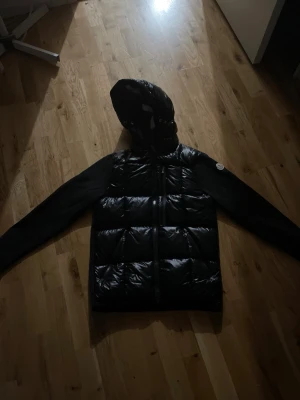 Moncler cardigan - Snygg svart dunjacka från Moncler med glansig, quiltad front och huva. Ärmarna är i ett mattare tyg och jackan har dragkedja framtill samt Moncler-logga på ena ärmen. Perfekt för kalla vinterdagar och riktigt stilren look.     Använt 1 gång ba kvitto finns nyttskick