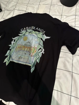 Svart Casablanca t-shirt med tryck - Snygg svart t-shirt från Casablanca med stort färgglatt tryck på ryggen som visar en arkad och bladmotiv. T-shirten har rund hals och korta ärmar. Perfekt för dig som vill sticka ut med en unik design.