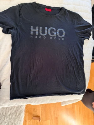 Svart HUGO BOSS t-shirt - Snygg svart t-shirt från HUGO BOSS med stor logga i silver på bröstet. Klassisk rund halsringning och korta ärmar. Perfekt för dig som gillar stilrena och trendiga plagg med tydlig branding.