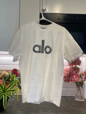 Vit t-shirt från Alo med logga - Snygg vit t-shirt från Alo med stor svart logga på bröstet. Klassisk rund halsringning och korta ärmar. Tillverkad i mjuk bomull för en skön känsla. Perfekt till jeans eller shorts för en clean och enkel stil.