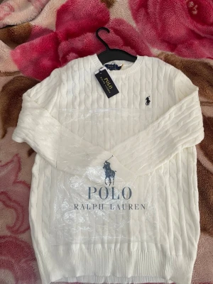 Vit kabelstickad tröja Polo Ralph Lauren - Kabelstickad vit tröja från Polo Ralph Lauren med rund halsringning och klassisk marinblå logga broderad på bröstet. Tröjan har ribbade muddar vid ärmslut och nederkant samt stor logotyp framtill. Perfekt för en stilren och tidlös look.
