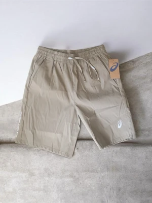 Beige shorts från Asics med snörning - Snygga beige shorts från Asics med elastisk midja och snörning. Tillverkade i lätt syntetmaterial som passar perfekt för träning eller varma dagar. Diskret Asics-logga på benet och praktiska sidofickor.