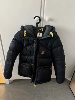 Svart pufferjacka från Fjällräven - Snygg svart pufferjacka från Fjällräven med huva och gula detaljer på snörningarna. Jackan har två stora fickor framtill med knappar och ett klassiskt Fjällräven-märke på bröstet. Perfekt för kalla vinterdagar och riktigt varm. Fint skick, inga skador eller så. 