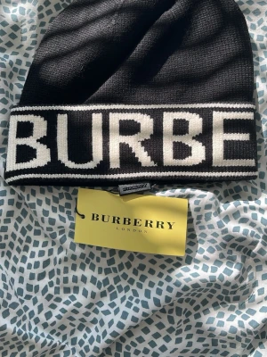 Svart Burberry mössa med logga - Snygg svart mössa från Burberry med stor vit logga runt om. Tillverkad i mjukt stickat material som håller dig varm. Klassisk och stilren design som passar perfekt till höstens och vinterns outfits.