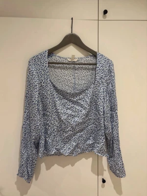 Blommig blå långärmad blus - Supersöt blus med blått och vitt småblommigt mönster och draperad urringning. Långärmad modell med lös passform och lätt, mjukt tyg som känns skönt mot huden. Perfekt för dig som gillar romantisk och trendig stil.