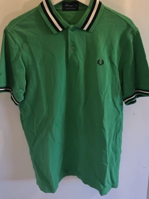 Grön pikétröja från Fred Perry - Klassisk grön pikétröja från Fred Perry med vit och svart rand på krage och ärmslut. Tröjan har korta ärmar, knappslå framtill och den ikoniska Fred Perry-loggan broderad på bröstet. Perfekt till en avslappnad och sportig stil.