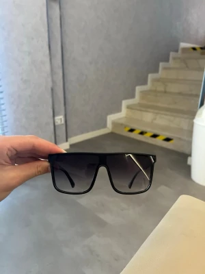 Svarta solglasögon från Emporio Armani - Säljer ett par svarta solglasögon från Emporio Armani med breda bågar och rektangulär form. Glasen är tonade och bågarna har en diskret logga på sidan. Perfekta för en clean och modern look.
