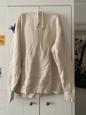 Off-White beige hoodie med tryck - Säljer en beige hoodie från Off-White med stor huva och magficka. På ryggen finns ett stort rött tryck med texten 'MERCY' i bubbliga bokstäver. Hoodien har ribbade muddar och en avslappnad passform. Perfekt för dig som gillar streetwear och unika detaljer.       (Kristet ”märke”)