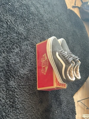 Grå Vans Old Skool med foder - Säljer ett par grå Vans Old Skool sneakers med vit sula och klassisk vit sidostripes. Skorna har ett mysigt beige foder i syntetpäls som gör dem extra sköna. Ovandelen är i mockaliknande material och de har snörning framtill. Perfekta för dig som vill ha både stil och komfort.