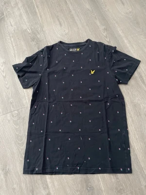 Svart t-shirt från Lyle & Scott - Svart t-shirt från Lyle & Scott med små diskreta mönster över hela tröjan och den klassiska gula loggan broderad på bröstet. T-shirten har rund hals och korta ärmar, tillverkad i mjuk bomull för skön känsla.