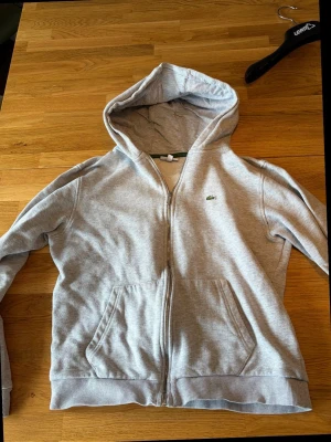 Grå zip hoodie från Lacoste - Snygg grå hoodie från Lacoste med dragkedja och klassisk krokodil-logga på bröstet. Hoodien har två stora fickor fram och ribbade muddar vid ärmar och nederkant. Perfekt för chill dagar och enkel att matcha med allt. Materialet är mjukt och skönt bomullstyg.aldrig använd
