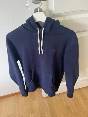Mörkblå hoodie från Polo Ralph Lauren - Snygg mörkblå hoodie från Polo Ralph Lauren med klassisk huva och vita dragsnören. Tröjan har en broderad röd logga på bröstet och en stor magficka framtill. Perfekt för en avslappnad och stilren look. Nypris 2500