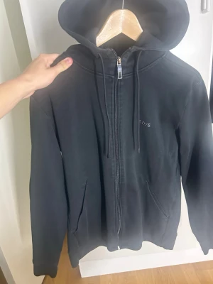 Svart zip hoodie från BOSS - Svart hoodie från BOSS med dragkedja framtill, två fickor och justerbar huva med snören. Diskret BOSS-logga på bröstet och gröna detaljer på insidan av kragen. Tillverkad i mjukt bomullsmaterial för en skön känsla.