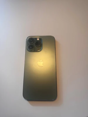 iPhone 13 Pro alpine green - Snygg iPhone 13 Pro i alpine green färg med stilren design och mattslipad baksida i glas. Telefonen har tre stora kameralinser och den klassiska Apple-loggan på baksidan. Ramen är i metall och framsidan har en stor, svart skärm.128 gb lagringsutrymme och 77% batteri hälsa