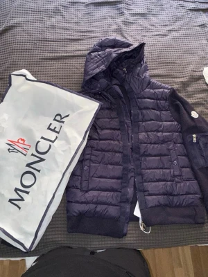 Mörkblå dunväst från Moncler - Snygg mörkblå dunväst från Moncler med huva och dragkedja framtill. Västen har två fickor med knappar och ribbade muddar. Klassisk Moncler-logga på ärmen och coolt serietryck på insidan. Perfekt för lager-på-lager och streetstyle.