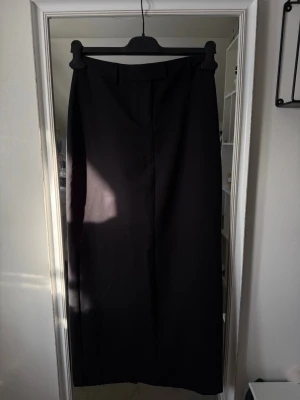 Svart långkjol med medelhög midja - Klassisk svart långkjol med medelhög midja och bälteshällor. Kjolen har en rak och stilren siluett som passar till många olika looks. Tillverkad i ett mjukt och följsamt material som ger en snygg passform.