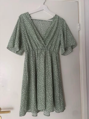 Grön v-ringad klänning - Söt grön kortklänning med vitt blommönster och omlott framtill. Klänningen har korta, vida ärmar och resår i midjan för en bekväm passform. Perfekt för vår och sommar!