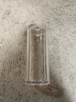 Transparent bomullspadsbehållare - Praktisk och stilren behållare för bomullspads i transparent plast. Cylindrisk form med öppning framtill och lock med rund knopp för enkel hantering. Perfekt för att hålla ordning på sminkrutinens tillbehör på sminkbordet eller i badrummet.