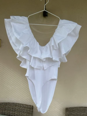  Ny  Trendig Vit baddräkt med volanger - Trendig vit baddräkt med djup urringning och stora volanger längs axelbanden. Baddräkten har en feminin och iögonfallande design, perfekt för dig som vill sticka ut på stranden eller vid poolen. Materialet ser ut att vara stretchigt och bekvämt.