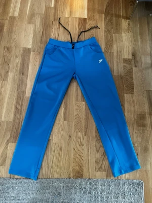 Blå Nike mjukisbyxor med raka ben - Snygga blå mjukisbyxor från Nike med raka ben och dragsko i midjan. Byxorna har två sidofickor och en liten vit Nike-logga på vänster sida. Perfekta för chill eller träning. Materialet känns mjukt och stretchigt.