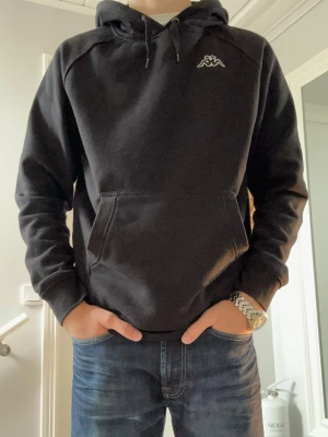 Svart hoodie från Kappa - Snygg svart hoodie från Kappa med klassisk logga broderad på bröstet. Hoodien har en stor huva med dragsko och en rymlig magficka. Tillverkad i mjukt material som känns skönt mot huden. Perfekt för en avslappnad och sportig stil. Storlek: L Modellen är 181cm. 