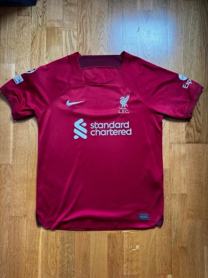 Liverpool matchtröja Nike röd - Snygg röd Liverpool matchtröja från Nike med klubbmärke och sponsortryck på bröstet. Tröjan har korta ärmar, rund hals och Champions League-märken på ärmarna. Tillverkad i lätt och ventilerande funktionsmaterial, perfekt för fotboll eller träning.