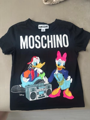 Svart Moschino x H&M Disney T-shirt - Cool svart T-shirt från Moschino x H&M med stort vitt MOSCHINO-tryck och färgglad print av Kalle Anka och Kajsa Anka i streetwear-stil. T-shirten har rund hals och korta ärmar. Perfekt för dig som gillar statement-plagg och Disney.