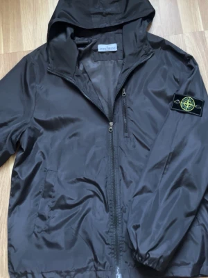 Stone Island windbreaker - Snygg svart vindjacka från Stone Island med huva och dragkedja framtill. Jackan har två sidofickor och den klassiska Stone Island-loggan på vänster ärm. Tillverkad i ett lätt, glansigt material som passar perfekt för blåsiga dagar. OBS!! kolla sista bilden.
