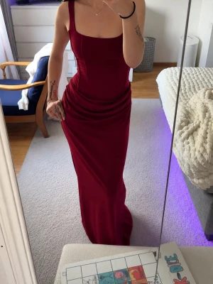 Vinröd balklänning - Säljer en elegant vinröd långklänning med figurnära passform och breda axelband. Klänningen har snygg snörning i ryggen som ger en justerbar och unik look. Perfekt för speciella tillfällen när du vill sticka ut.