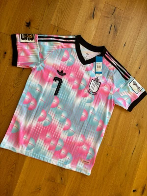 Adidas retro fotbollströja rosa/blå - Vm fotbollströja från Adidas med färgstarkt mönster i rosa, blått och vitt. Svart V-ringad krage och svarta detaljer på ärmarna. Nummer 7 på bröstet och klassiska Adidas-ränder på axlarna. Tillverkad i lätt och ventilerande material. Oanvänd 