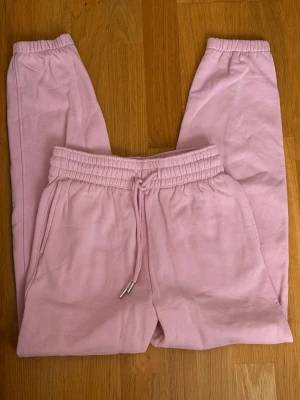 Rosa mjukisbyxor från H&M - Säljer ett par ljusrosa mjukisbyxor från H&M med dragsko i midjan och resår vid bensluten. Byxorna har två sidofickor och är gjorda i en mjuk bomulls- och polyesterblandning. Perfekta för chill dagar eller när du vill vara extra bekväm.