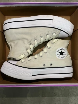 Vita Converse Chuck Taylor Highs - Klassiska vita Converse Chuck Taylor All Star high top sneakers i canvas med vita snören och svart rand längs sulan. Ikonisk logga på sidan och tåhätta i gummi. Perfekta för en avslappnad och trendig stil. Flexibel med priset