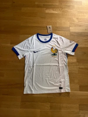 Frankrike fotbollströja Nike vit - Snygg vit fotbollströja från Nike med tunna blå och röda ränder. Tröjan har blå krage och ärmslut samt Frankrikes tuppemblem med två stjärnor broderat på bröstet. Perfekt för dig som älskar fotboll och vill stötta Frankrike.   Fraktar samma eller dagen efter du köpt.