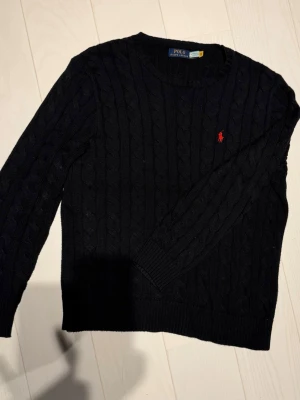 Svart kabelstickad tröja Polo Ralph Lauren - Kabelstickad svart tröja från Polo Ralph Lauren med röd broderad logga på bröstet. Rund halsringning, långa ärmar och ribbade muddar. Klassisk och stilren design som passar till jeans eller chinos.