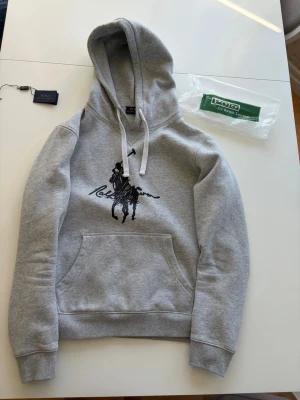 Grå hoodie från Polo Ralph Lauren - Snygg grå hoodie från Polo Ralph Lauren. Hoodien har huva med vita snören och en klassisk magficka. Materialet är mjukt och bekvämt, perfekt för en avslappnad stil. Knappt använd, säljer för den är för liten. Storlek L men passar M. Påse och etiketterna kommer med. Pris 699.