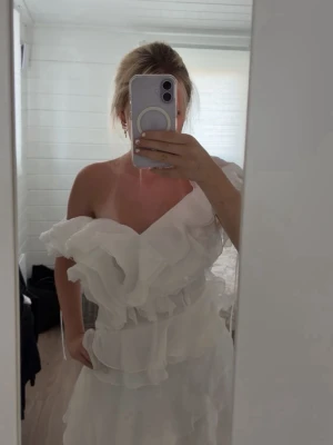 Vit volangklänning  - Supersöt vit klänning med volanger och oneshoulder-design. Klänningen har flera lager volanger som ger en romantisk och luftig känsla. Perfekt för sommarens alla tillfällen och har en unik axelbandslös look.