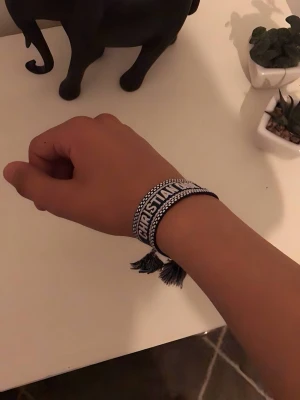 Dior armband - Säljer ett snyggt blått armband från Christian Dior med broderad logotyp och vita detaljer. Armbandet har justerbar knytning med tofsar och är gjort i textilmaterial. Perfekt för dig som vill ha en trendig accessoar med lyxig känsla.