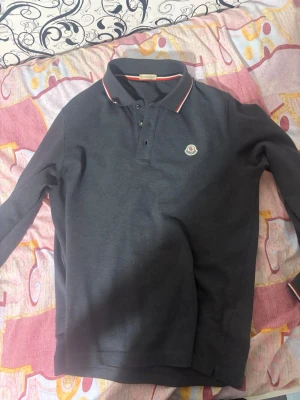 Mörkblå långärmad piké från Moncler - Snygg mörkblå långärmad pikétröja från Moncler med klassisk krage och knappar framtill. Diskret Moncler-logga på bröstet och rödvit detalj på kragen och ärmsluten. Perfekt för en stilren och avslappnad look.