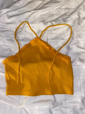 Gul/orange ribbad crop top med volang - Fläcken går bort i tvätt innan frakt! Säljer en gul/orange crop top med tunna axelband och volangdetalj längs halsringningen. Toppen är ribbad och har en figurnära passform, perfekt för sommaren. Materialet känns mjukt och stretchigt. 