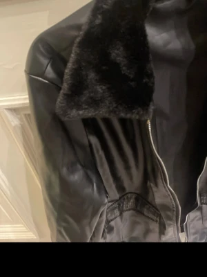 Svart skinnjacka med fuskpäls - Snygg svart skinnjacka med bred krage i fuskpäls och dragkedja framtill. Jackan har ribbade muddar vid ärmslut och nederkant samt två fickor på sidorna. Perfekt för dig som vill ha en klassisk men trendig look.
