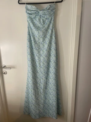 Ljusblå axelbandslös långklänning - Säljer en ljusblå axelbandslös långklänning med blommigt mönster. Klänningen har en v-ringad överdel med twistad detalj framtill. Perfekt för midsommar. Använd en gång och kosta 700 kr nypris. 