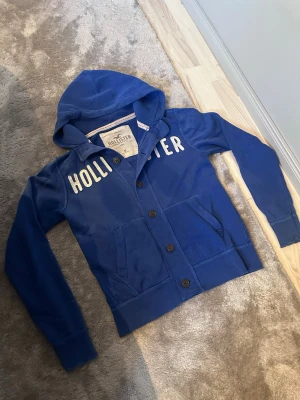 Vintage Hollister hoodie med knappar - Snygg och unik vintage hoodie från Hollister med stora vita bokstäver över bröstet. Hoodien har knäppning med bruna knappar, två fickor framtill och en rejäl huva. Tillverkad i mjukt bomullsmaterial som känns skönt mot huden. 