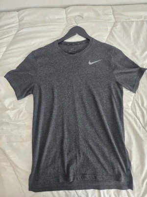 Mörkgrå Nike Dri-Fit t-shirt - Nyskick Snygg mörkgrå t-shirt från Nike med Dri-Fit-teknologi och klassisk rund hals. T-shirten har korta ärmar och en diskret Nike-logga på bröstet. Perfekt för träning eller chill dagar.