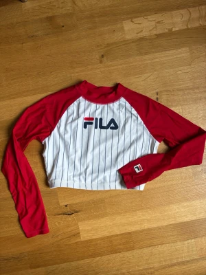 Vintage surf / croppad topp från FILA - Sportig croppad topp från FILA med vita och tunna marinblå ränder, röda ärmar och logga framtill. Långärmad modell med kontrastfärgade raglanärmar och liten logga på ärmen. Perfekt för en trendig och avslappnad look alternativt för att se snygg ut på surfbrädan. Strl M men liten i storleken, sitter mer som en S/XS
