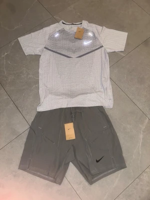 Nike träningsset t-shirt och shorts - Nike träningsset med en vit t-shirt i meshmaterial och gråa shorts. T-shirten har ett diskret mönster och reflekterande Nike-logga på bröstet. Shortsen är gråa med dragsko i midjan och svart Nike-logga på benet. Perfekt för gym eller löpning.